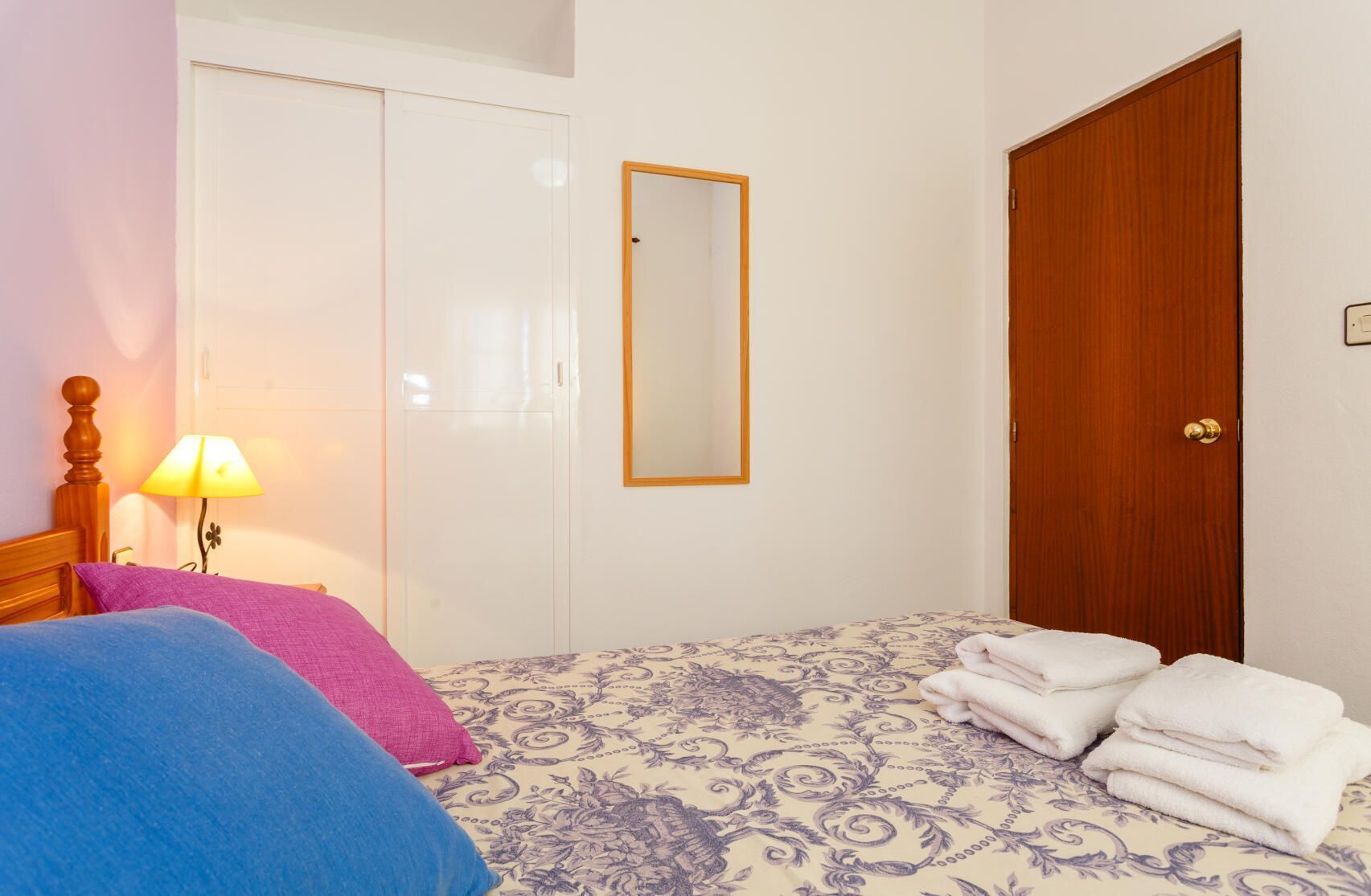 A bedroom with a bed, a bedside lamp, a mirror, a wardrobe, and a door.