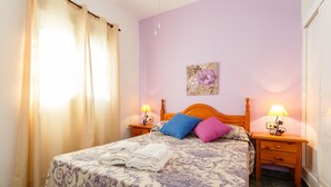 Ein ordentlich gemachtes Bett mit einem blumigen Bettbezug, zwei Kissen und einem hölzernen Kopfteil. Es gibt zwei Nachttische mit Lampen, ein an der Wand befestigtes Bild und ein Fenster mit Vorhängen.