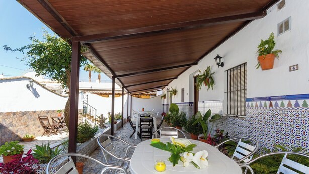 Un patio al aire libre con una mesa redonda y sillas, rodeado de plantas en macetas y una pared con azulejos.