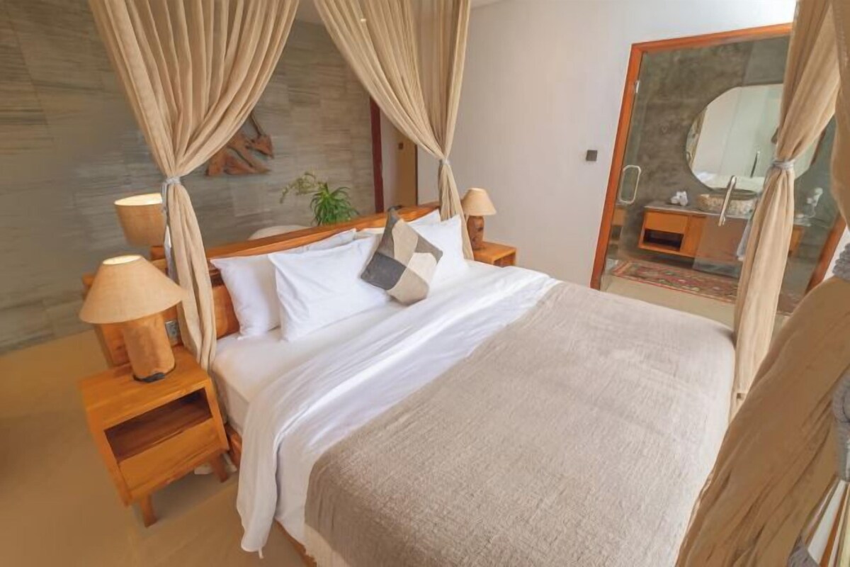 Een slaapkamer met een bed, houten nachtkastjes, een lamp en een glazen deur naar een badkamer.