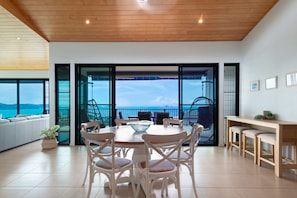 A modern dining area with a round table, white chairs, and a view of the sea through large glass doors.