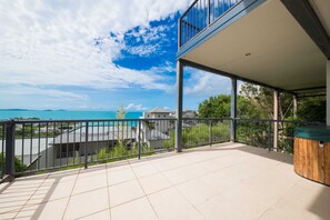 A balcony with a view of the sea and buildings, a wooden bench, and a tiled floor.