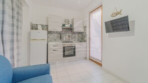 A compact kitchen with white cabinets, a stove, and a microwave. There is a blue sofa in the foreground and a fish decoration on the wall. - APPARTAMENTI LA MARINA - APPARTAMENTI LA MARINA - LIMONI (Marciana Marina)