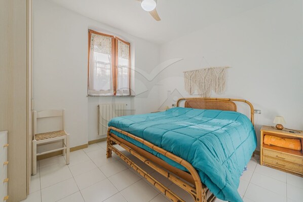 A bedroom with a wooden bed, a chair, a nightstand, a lamp, a window with curtains, and a wall-mounted macrame piece.