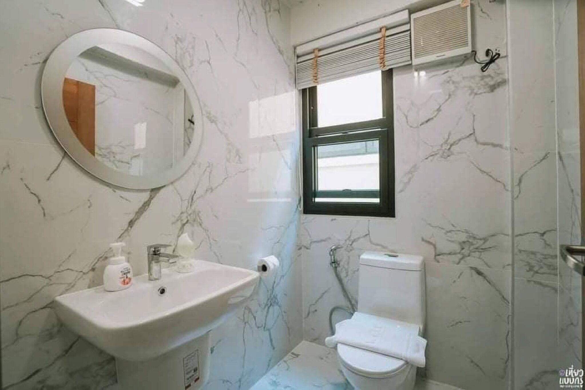 A bathroom with a marble wall, a round mirror, a sink, a toilet, and a window with blinds.