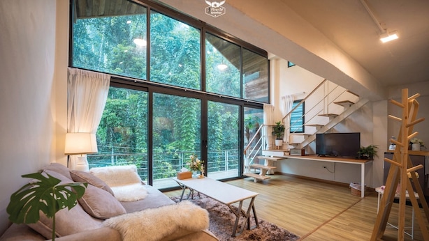 A modern living room with a large window, a sofa, a coffee table, and a staircase leading to an upper level. - My Cottage Chiangmai (Mae On)