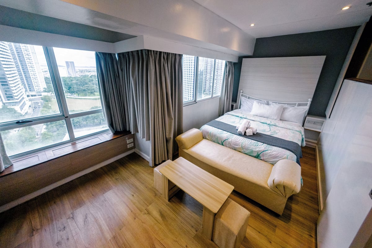 A modern hotel room with a large bed, a bench, a desk, and a view of the city through large windows.