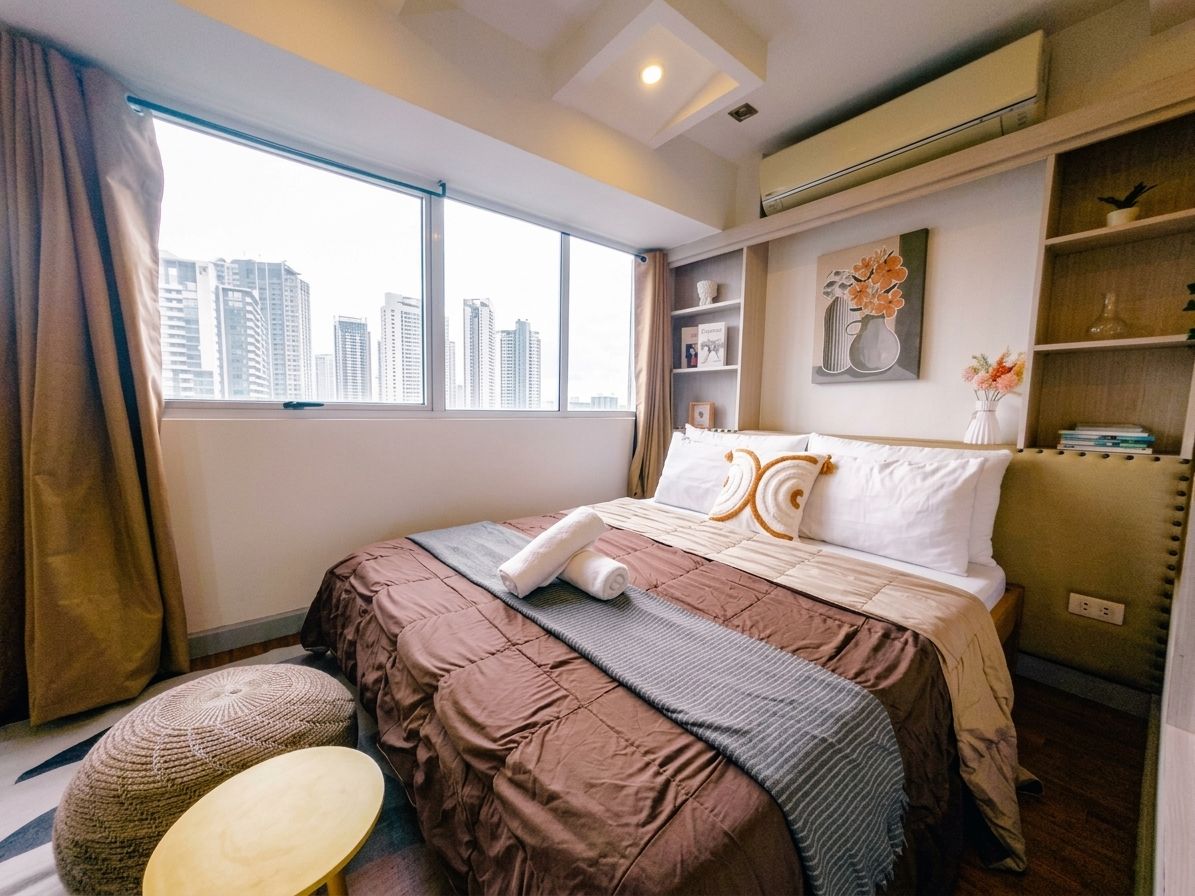 A hotel room with a large window, a bed with a brown and gray bedspread, a bedside table, and a small ottoman.