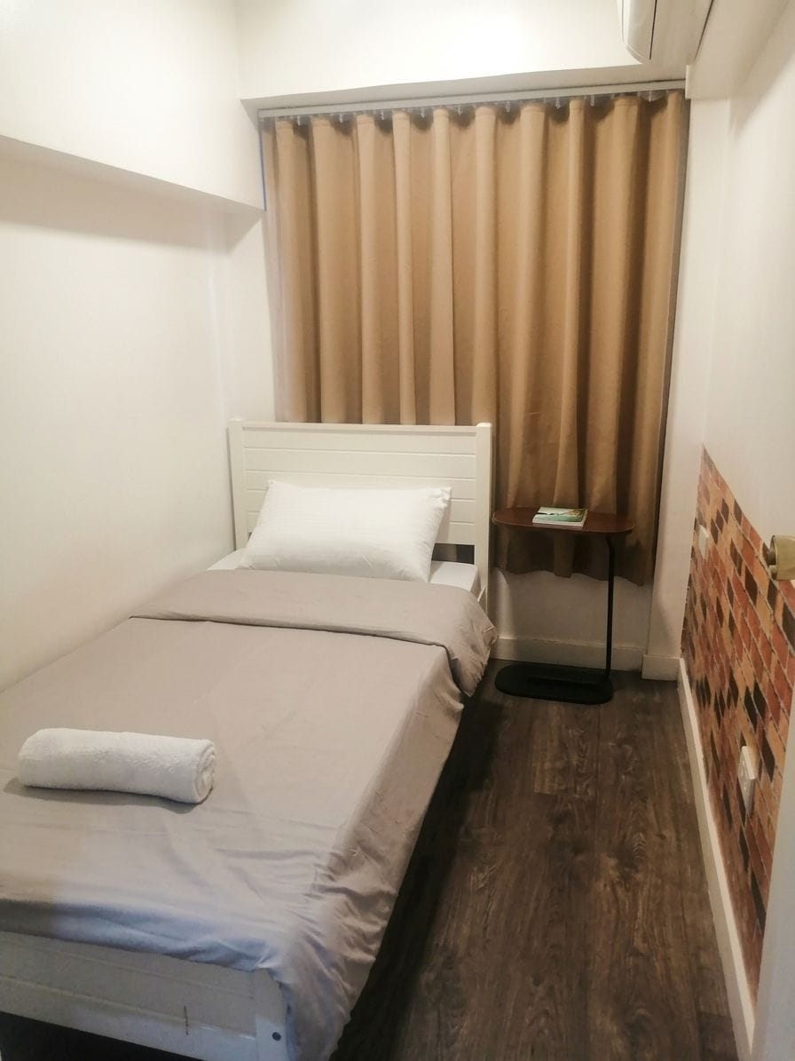A small, single-bed room with a wooden floor, a white headboard, and a window with curtains.