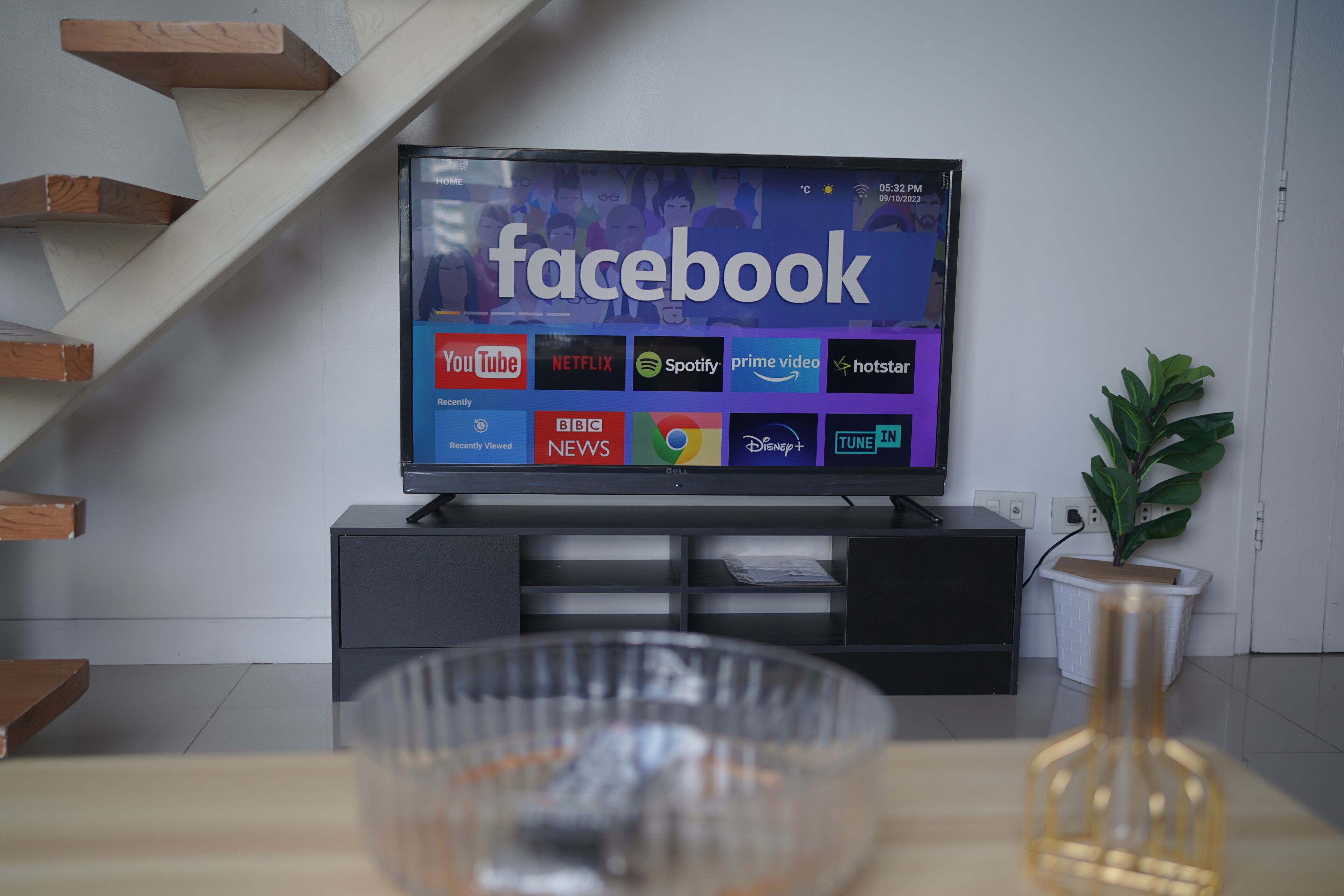 Ett modernt vardagsrum med en platt-TV som visar Facebook-appen, en trätrappa, en svart TV-möbel, en glasvas och en krukväxt.