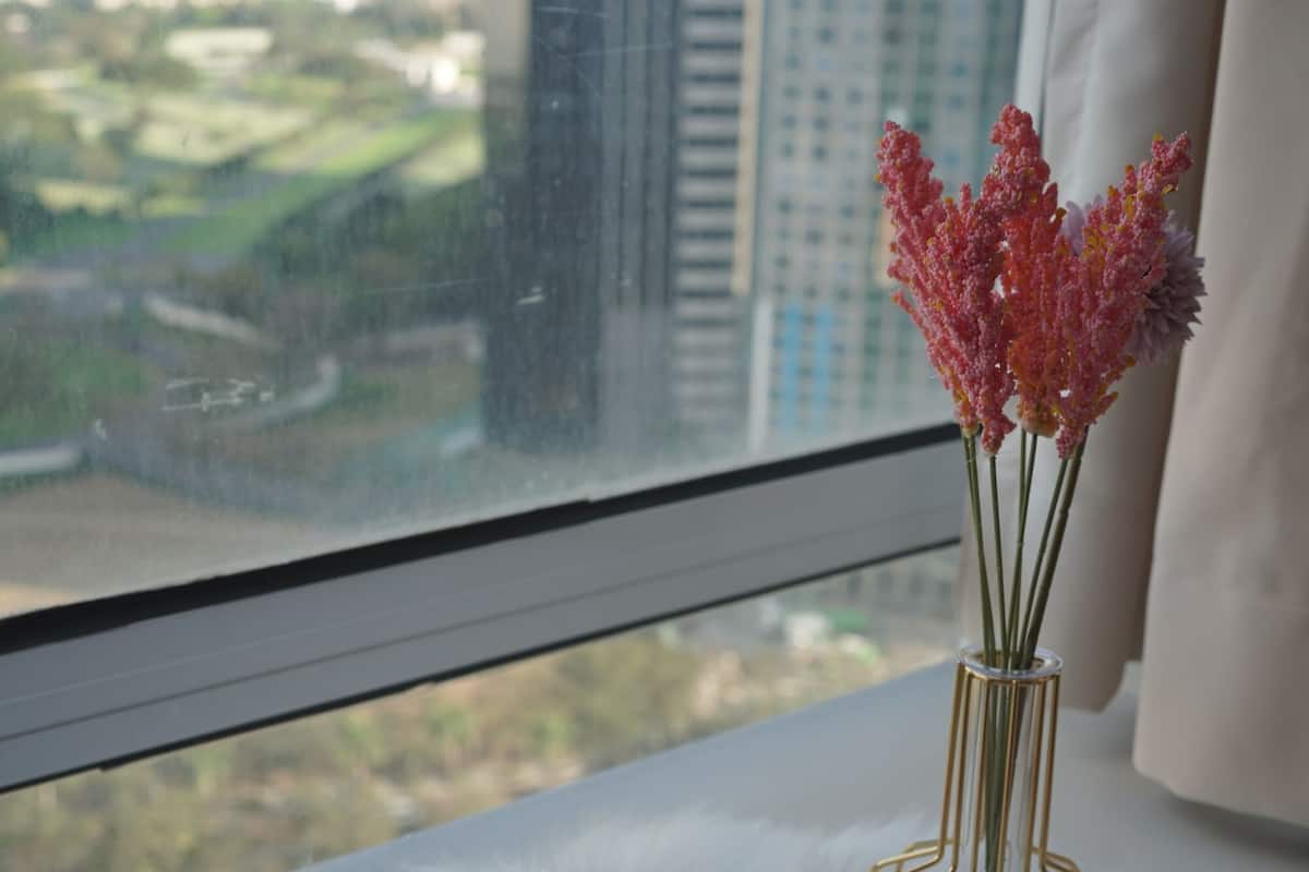 A pink hairdryer and a vase with pink flowers on a white furry surface by a window with a city view.