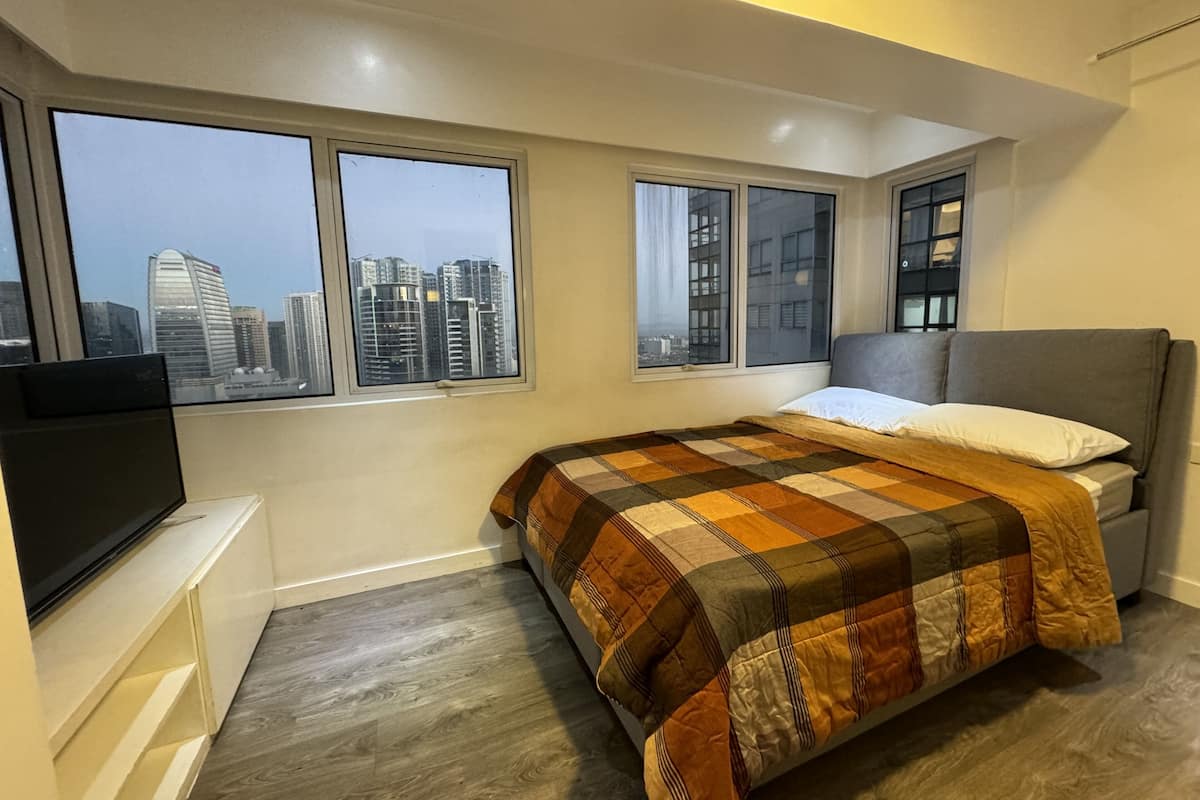 A bedroom with a bed, a TV, and large windows with a city view.