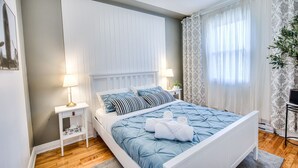 A bedroom with a white bed, blue bedding, a bedside table with a lamp, and a window with sheer curtains.