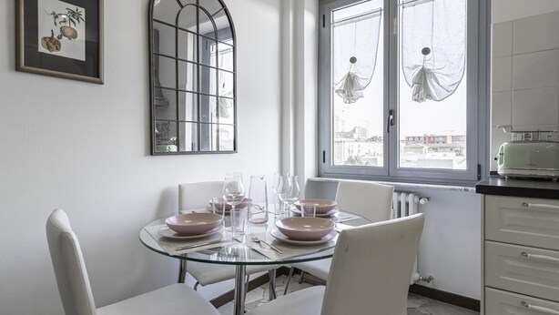 A dining area with a glass table set for four, white chairs, and a view of the city through a window with sheer curtains.