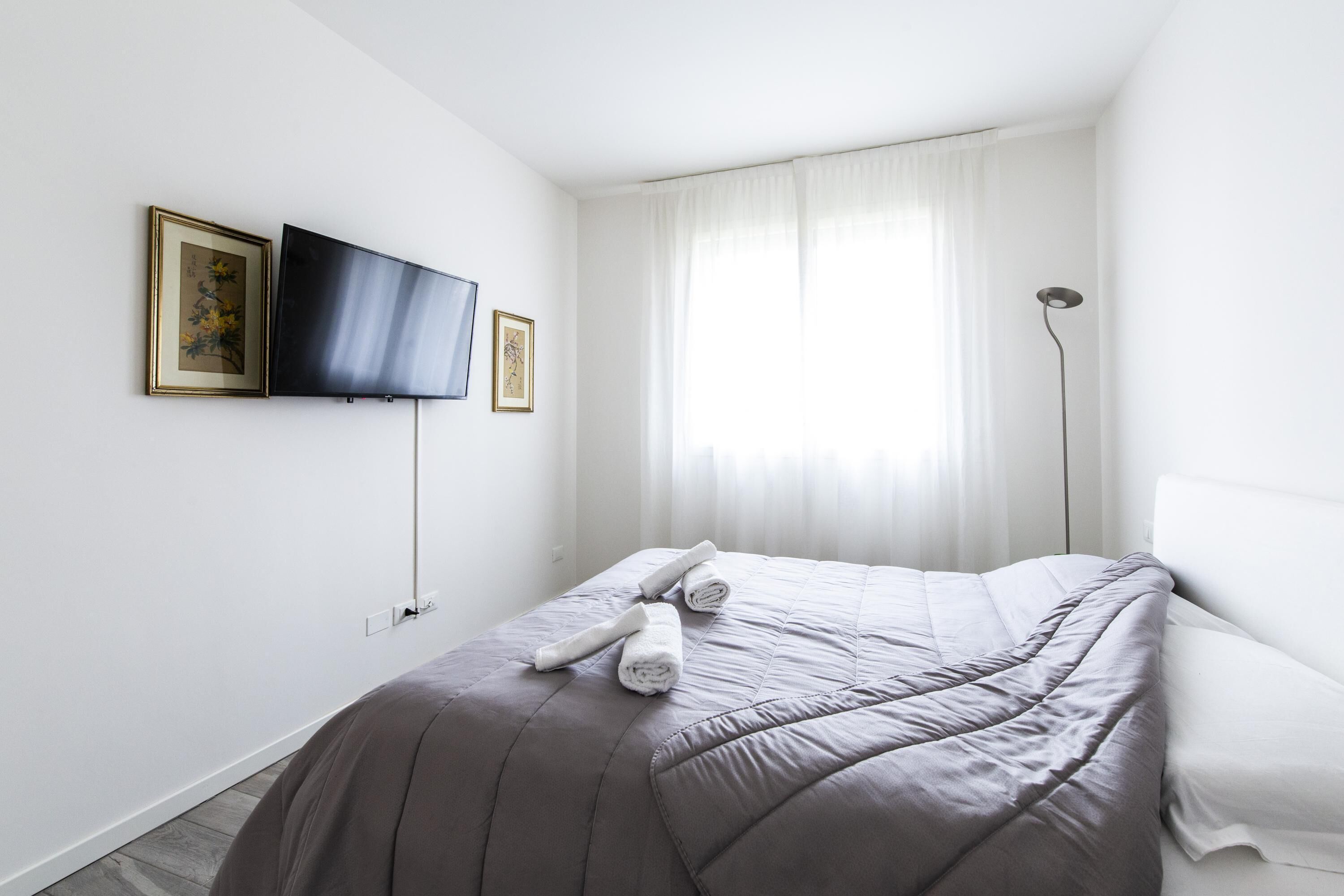 Ein ordentlich bezogenes Bett mit grauer Bettdecke, ein Flachbildfernseher an der Wand montiert und eine Stehlampe neben dem Bett.