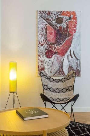 A room with a round wooden table, a woven chair, a wall-mounted painting, and a floor lamp.