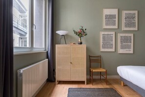 A room with a window, a radiator, a cabinet, a chair, a bed, and framed texts on the wall.