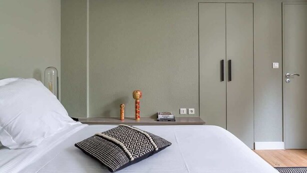 Een slaapkamer met een bed, kussens, een nachtkastje, een kledingkast en een klein tafeltje met een lamp en decoratieve spullen.