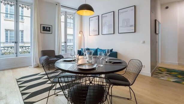 Un coin repas avec une table ronde, des chaises en fil de fer noir et un luminaire suspendu noir. Des tableaux encadrés ornent le mur, et un canapé avec des coussins bleus est également présent.