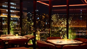 A restaurant with tables and chairs, large windows, and a view of the city at night.