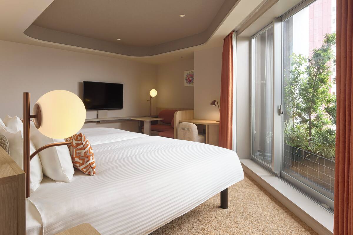 A hotel room with a large bed, a desk, a chair, a TV, and a balcony with a view of greenery.