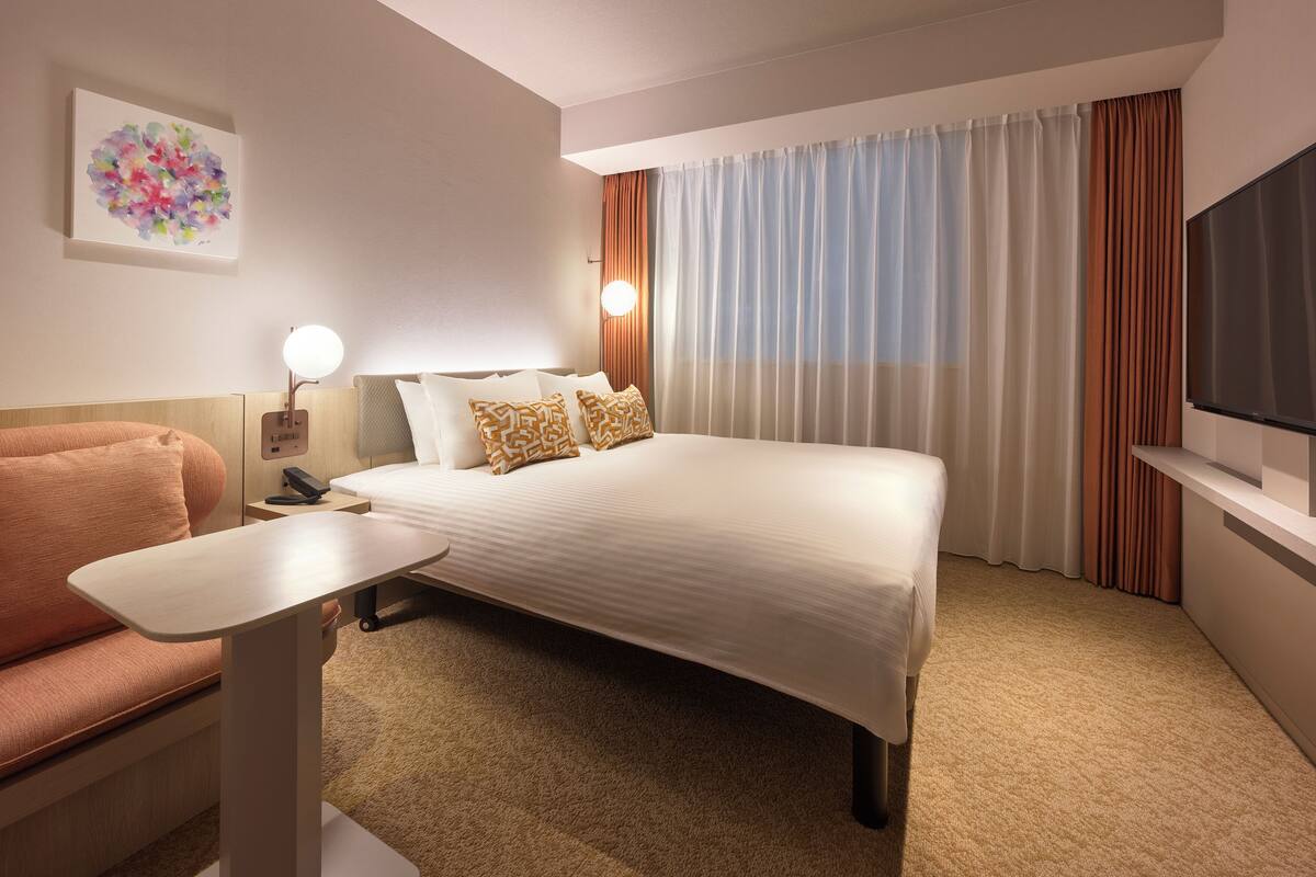 A hotel room with a bed, a desk, a chair, a TV, and a painting on the wall.