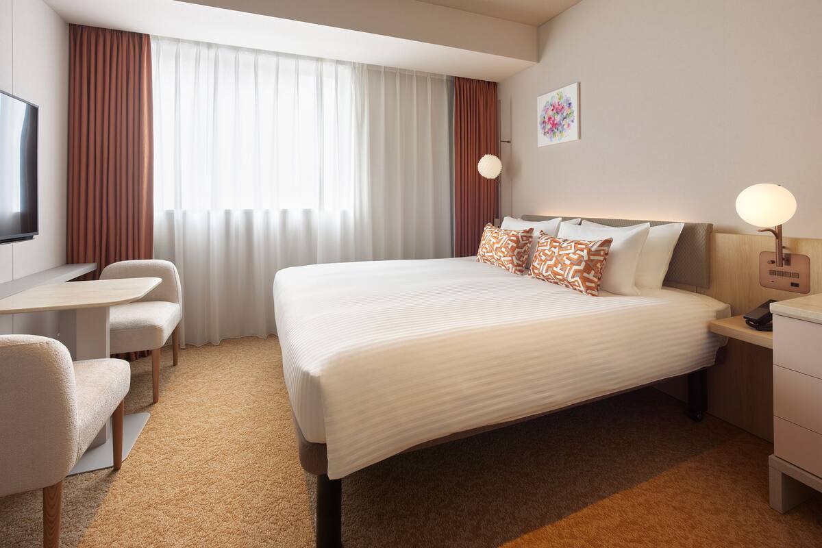 A hotel room with a bed, a desk, a chair, a nightstand, and a lamp.