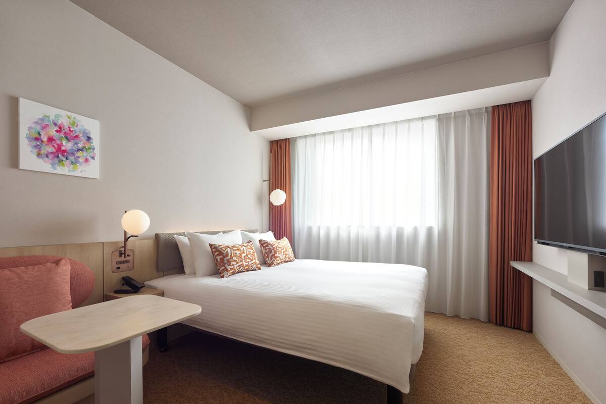 A hotel room with a bed, a desk, a chair, a TV, and a window with curtains.