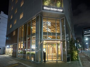 Exterior del Hotel Mitsui Garden de noche, con letreros iluminados, amplias ventanas de vidrio y una entrada visible.
