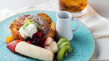 Een bord met Frans toast, opgeklopte room, aardbeien, bosbessen, plakjes banaan en kiwi, op een turquoise bord. Een witte mok en een glas ijsthee zijn ook zichtbaar.