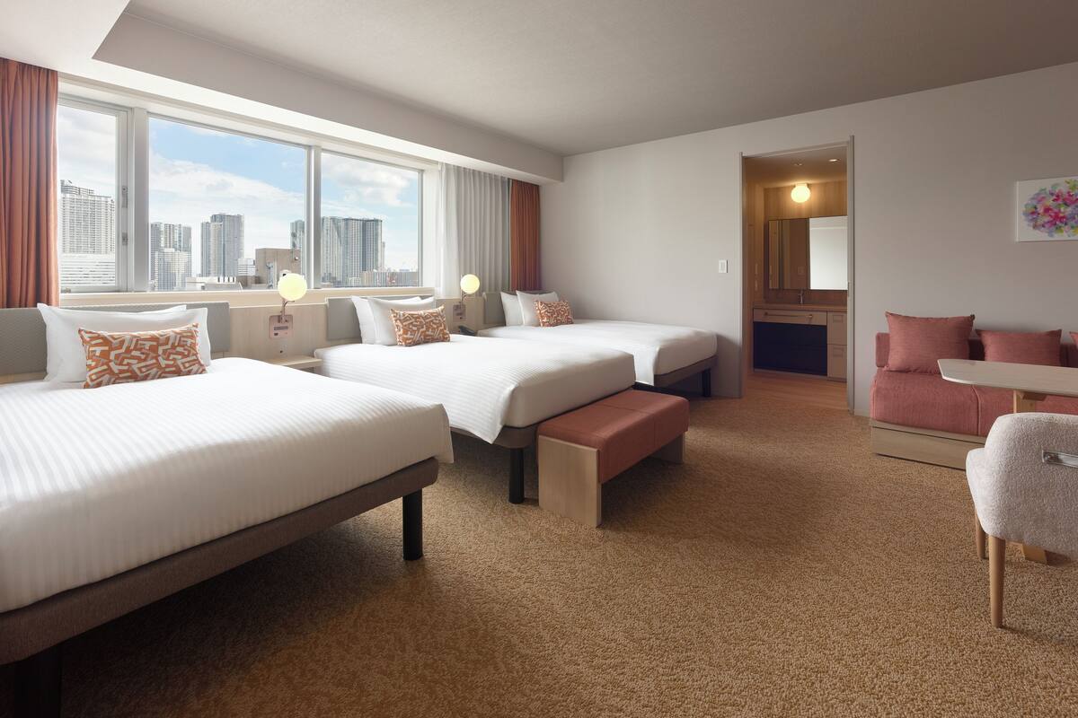 A hotel room with two beds, a bench, a sofa, a small table, and a view of the cityscape through large windows.