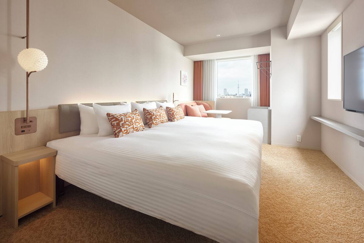 A modern hotel room with a large bed, a bedside table, a lamp, a TV, and a window with a city view.