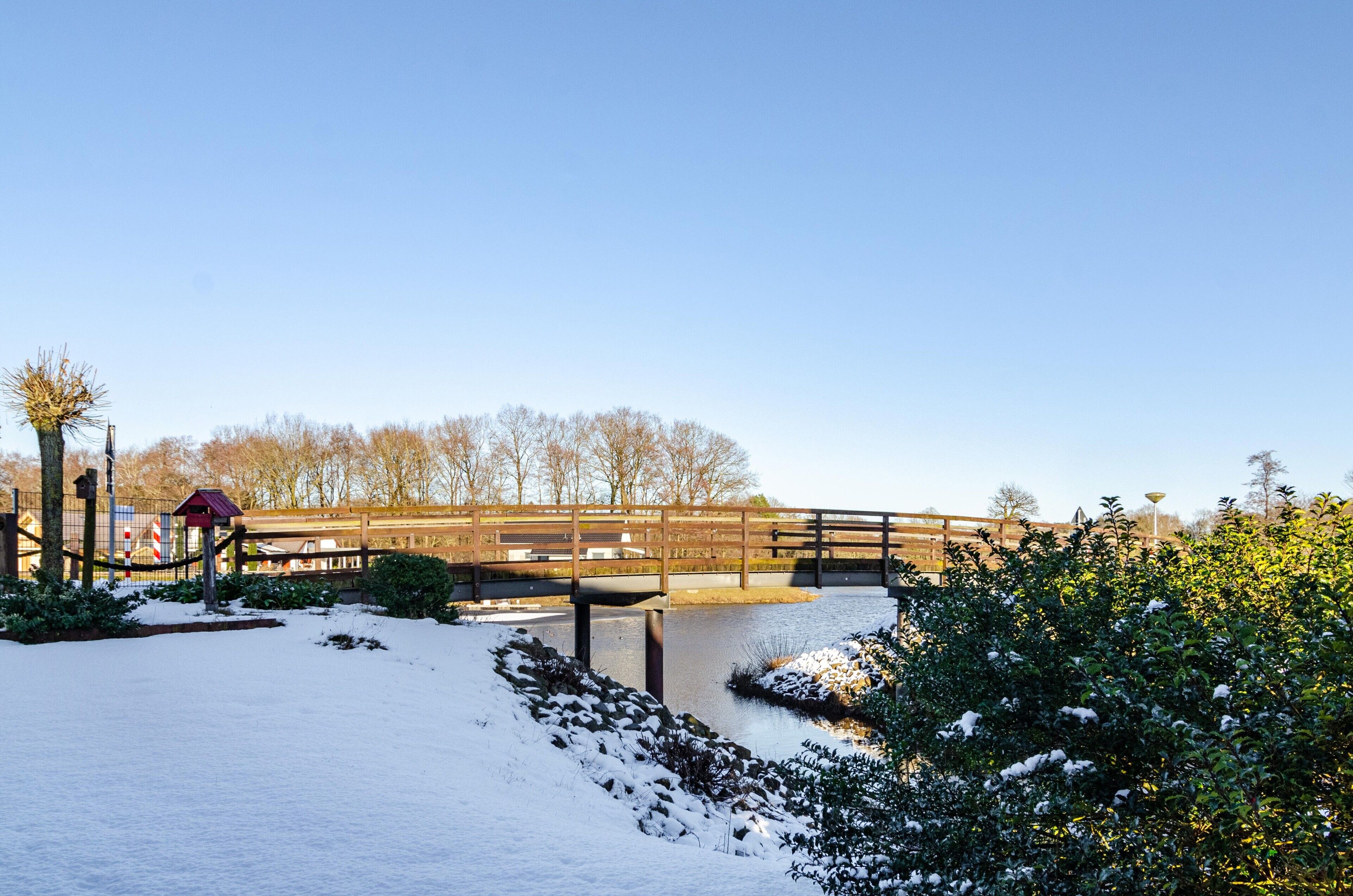 Eine Holzbrücke über einem verschneiten Fluss, umgeben von Bäumen und einem Zaun.