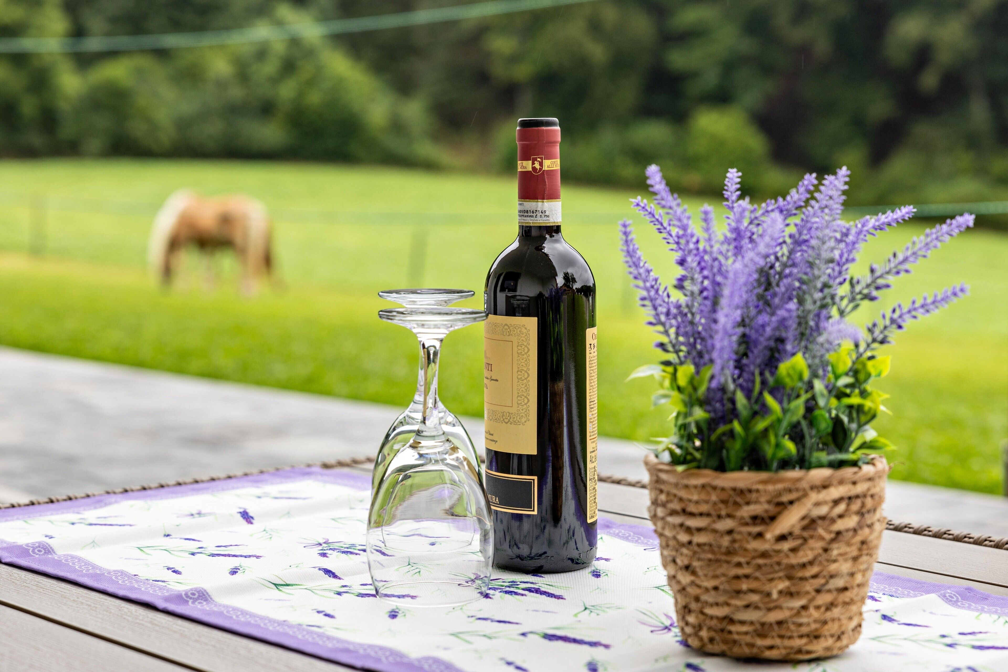 A bottle of wine, a wine glass, and a lavender plant on a table with a floral tablecloth.