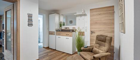 Un interior moderno con suelo de madera, un sillón de cuero marrón, un pequeño reposapiés, una zona de cocina con armarios blancos y un espejo.