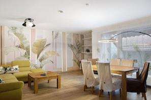 Una sala de estar con sofá, mesa de centro, mesa de comedor, sillas y un amplio ventanal con cortinas.