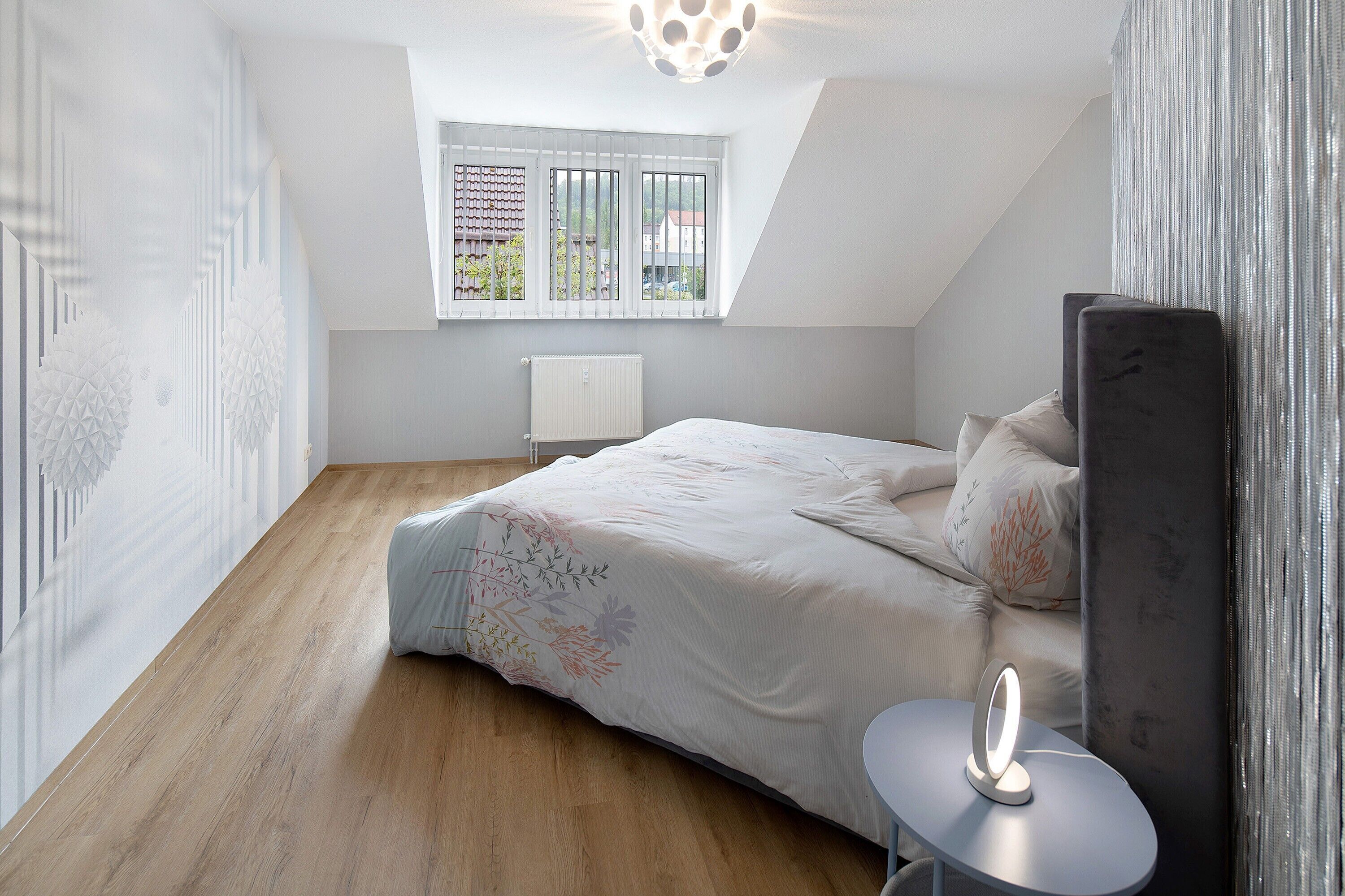A bedroom with a bed, a bedside table with a lamp, a window with blinds, and a radiator.