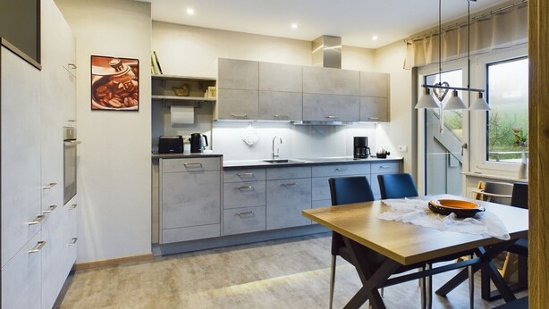 A modern kitchen with grey cabinets, a wooden dining table, and a view of the outdoors through a large window. - Apartment 'Wiesenblick' with Balcony and Wi-Fi (Brilon)