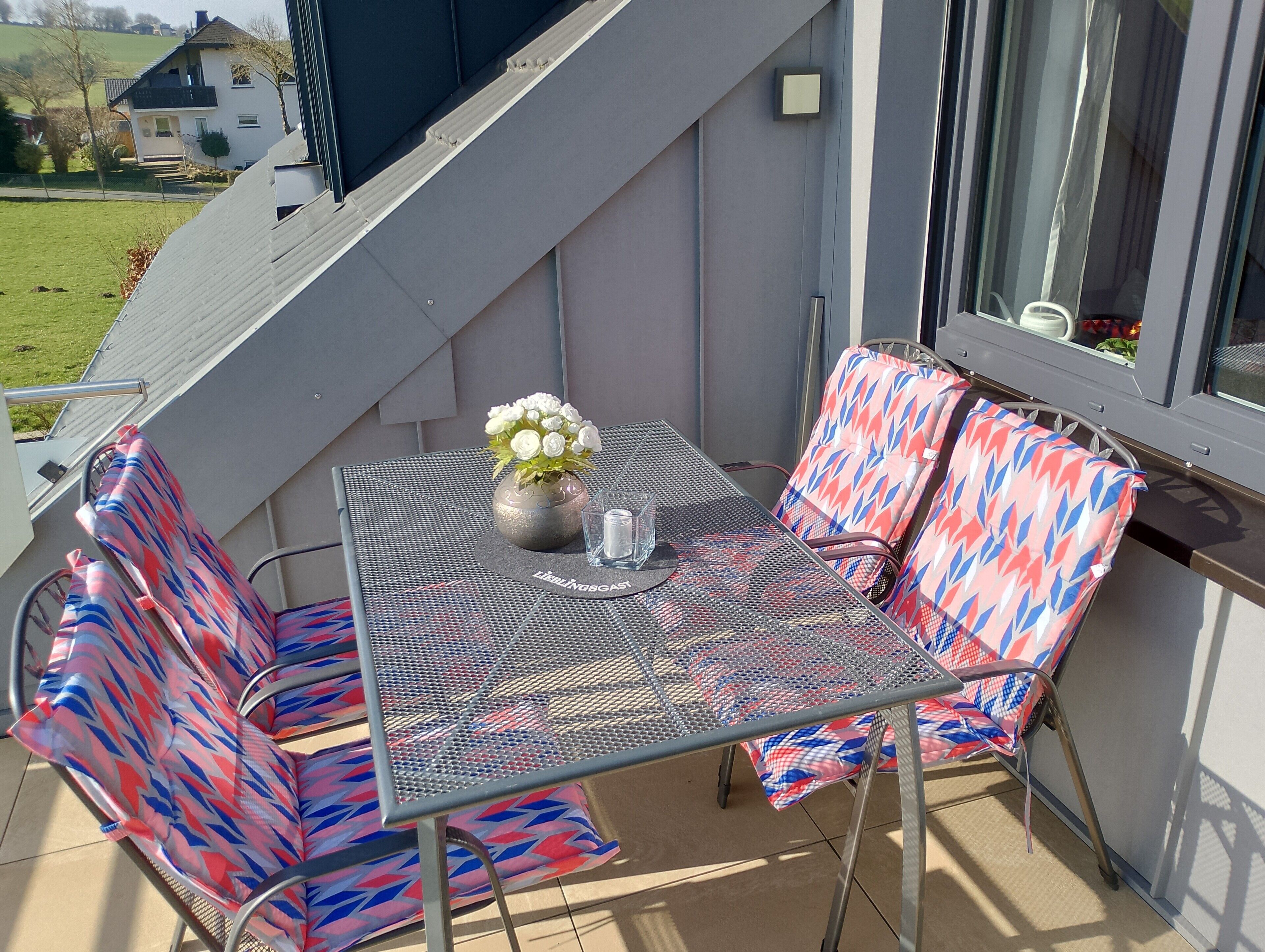 A balcony with a table, two chairs, a vase with flowers, and a window.