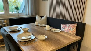 A dining table set with plates, bowls, and cutlery, a window with curtains, a black cushioned bench, and a patterned pillow. - Apartment 'Wiesenblick' with Balcony and Wi-Fi (Brilon)