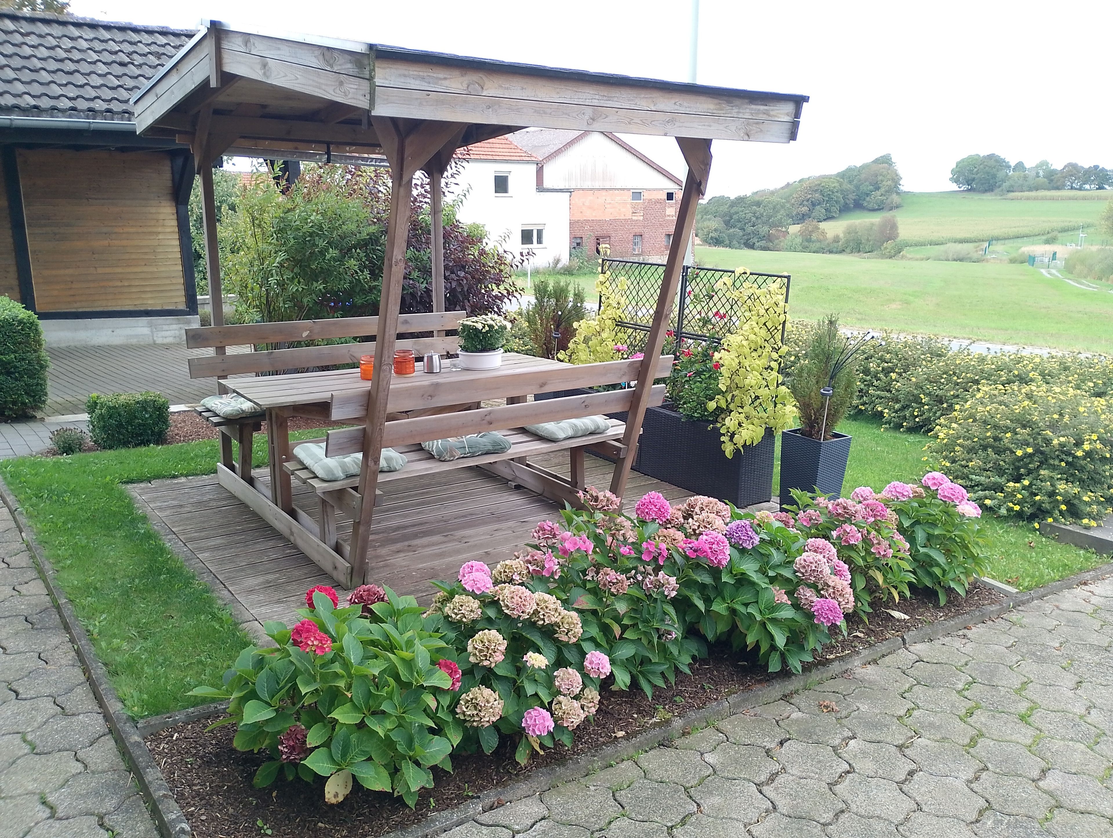 Une pergola en bois, agrémentée d’un banc et d’une table, entourée de parterres de fleurs roses et jaunes.