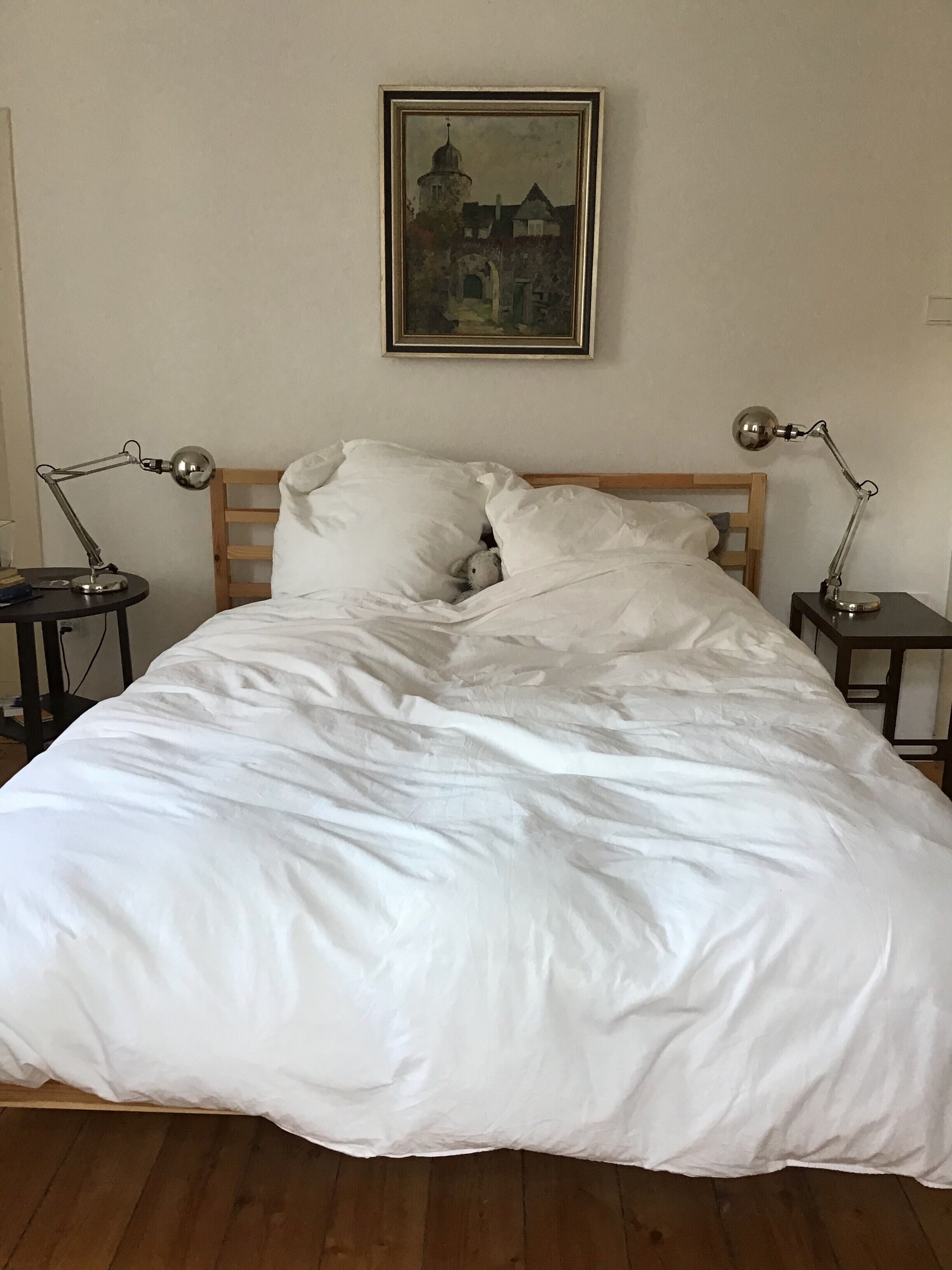 Ein ordentlich bezogenes Bett mit weißen Leinen, ein hölzernes Kopfteil, zwei Nachttische mit Lampen und ein gerahmtes Bild an der Wand.