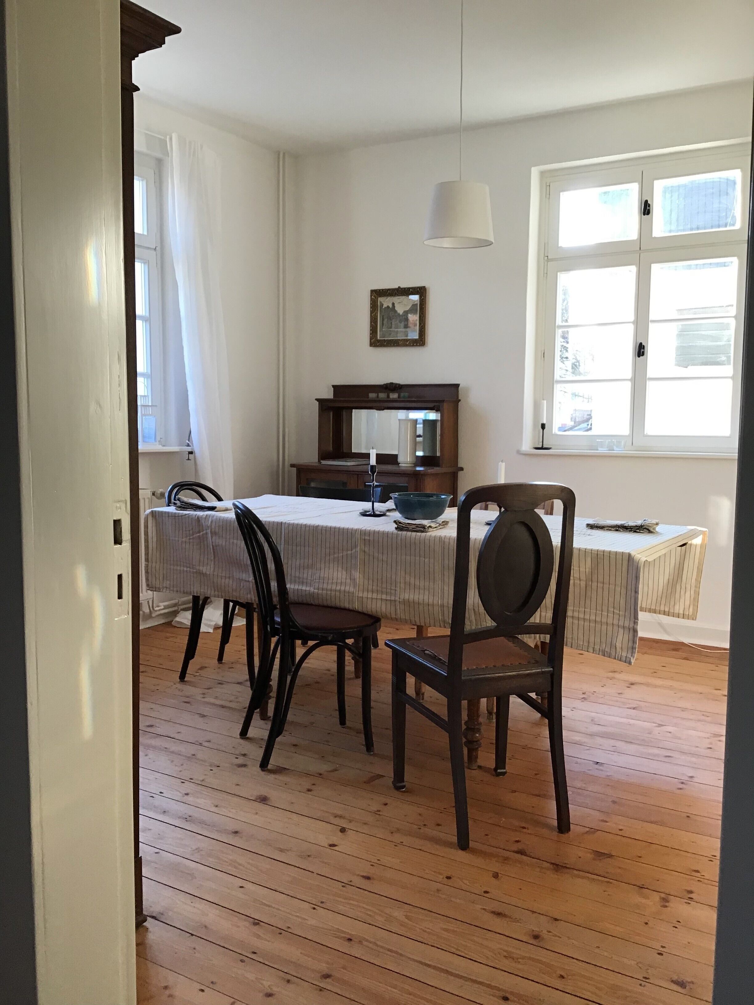 Un área de comedor con una mesa, sillas, un candelabro colgante, una ventana con cortinas y un cuadro en la pared.