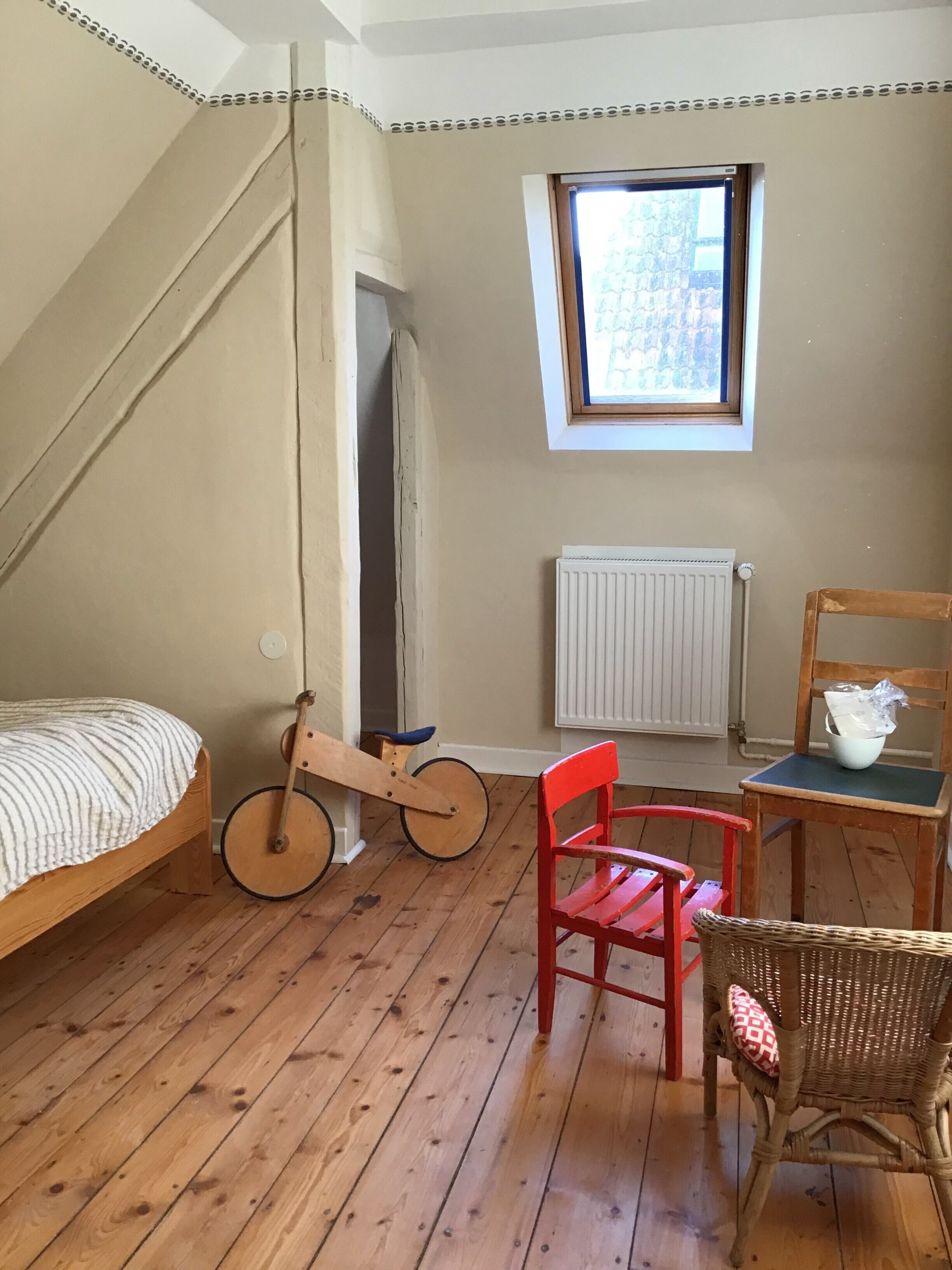 Une pièce comprenant un lit, un fauteuil à bascule en bois, une chaise rouge, une chaise en osier, une fenêtre et un radiateur.