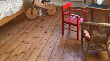 Une pièce comprenant un lit, un fauteuil à bascule en bois, une chaise rouge, une chaise en osier, une fenêtre et un radiateur.