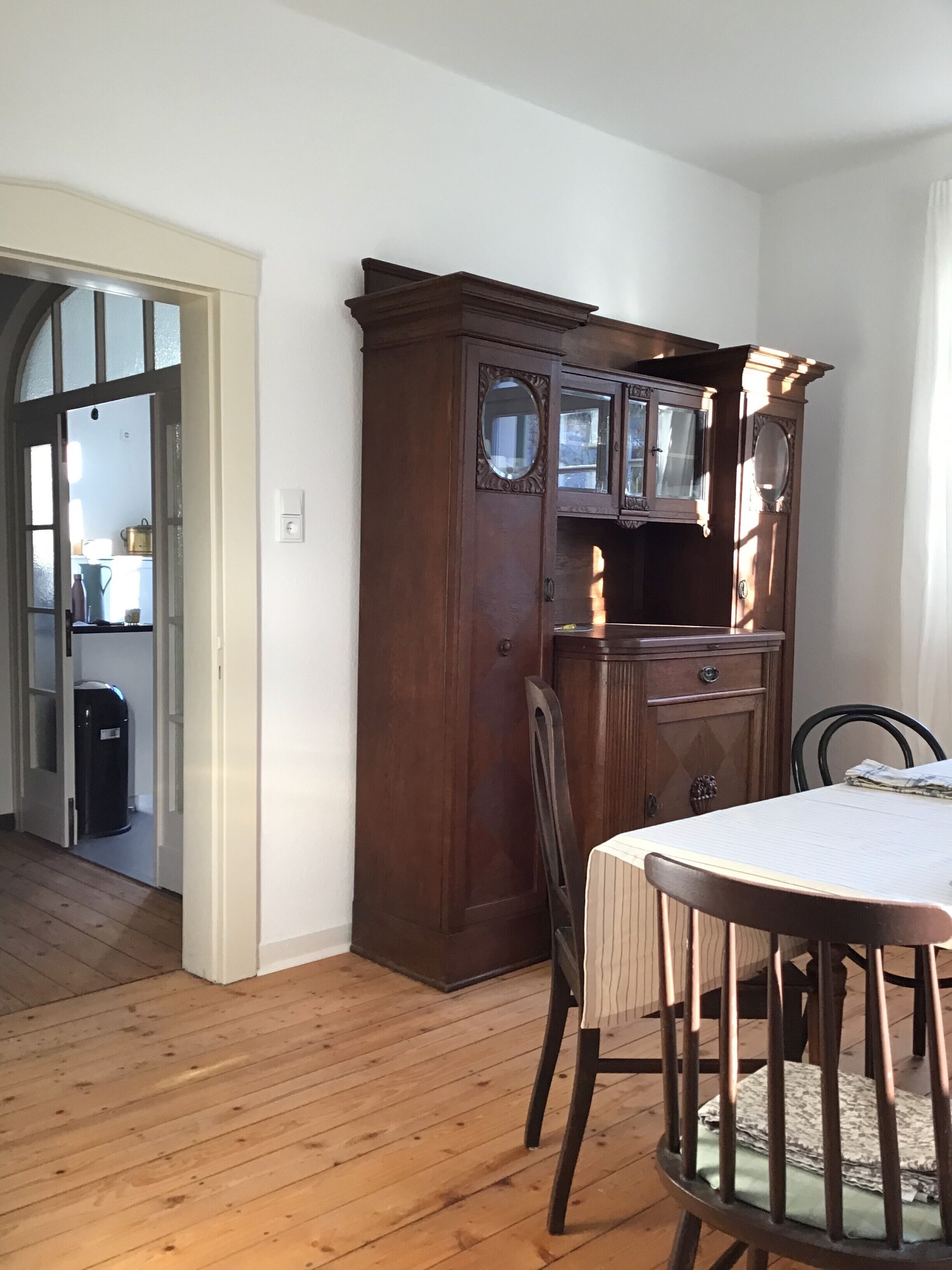 Un comedor con una mesa y sillas de madera, un mueble bajo de madera oscura con puertas de cristal, y un acceso a otra habitación.