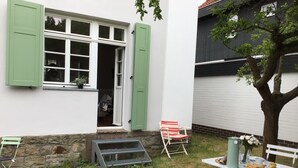 Una casa con cimientos de piedra, una ventana con contraventanas verdes, un pequeño porche con una silla y un árbol.
