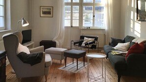 Una sala de estar con un sofá, un sillón, un reposapiés y una mesita. Hay una lámpara, un cuadro en la pared y una ventana con cortinas.