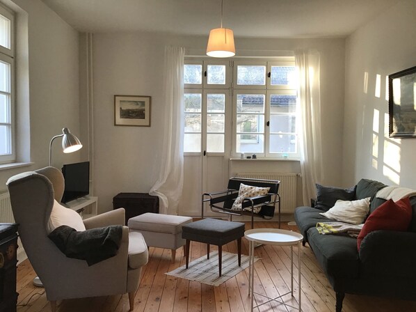 A living room with a sofa, armchair, ottoman, and a small table. There is a lamp, a picture on the wall, and a window with curtains.