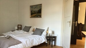 Un dormitorio con cama, almohadas, una mesita de noche con lámpara, un cuadro en la pared y una puerta que da a otra habitación.